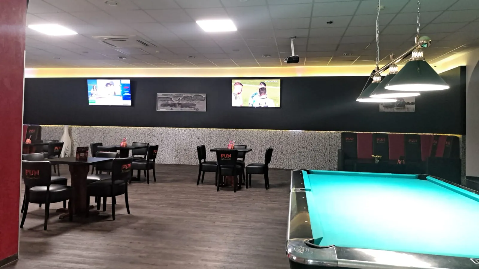 Gastraum von Billard Wesel mit Pooltisch unter grünen Lampen, an der schwarzen Wand zwei Fernseher mit Sport, davor Tische und Lederstühle