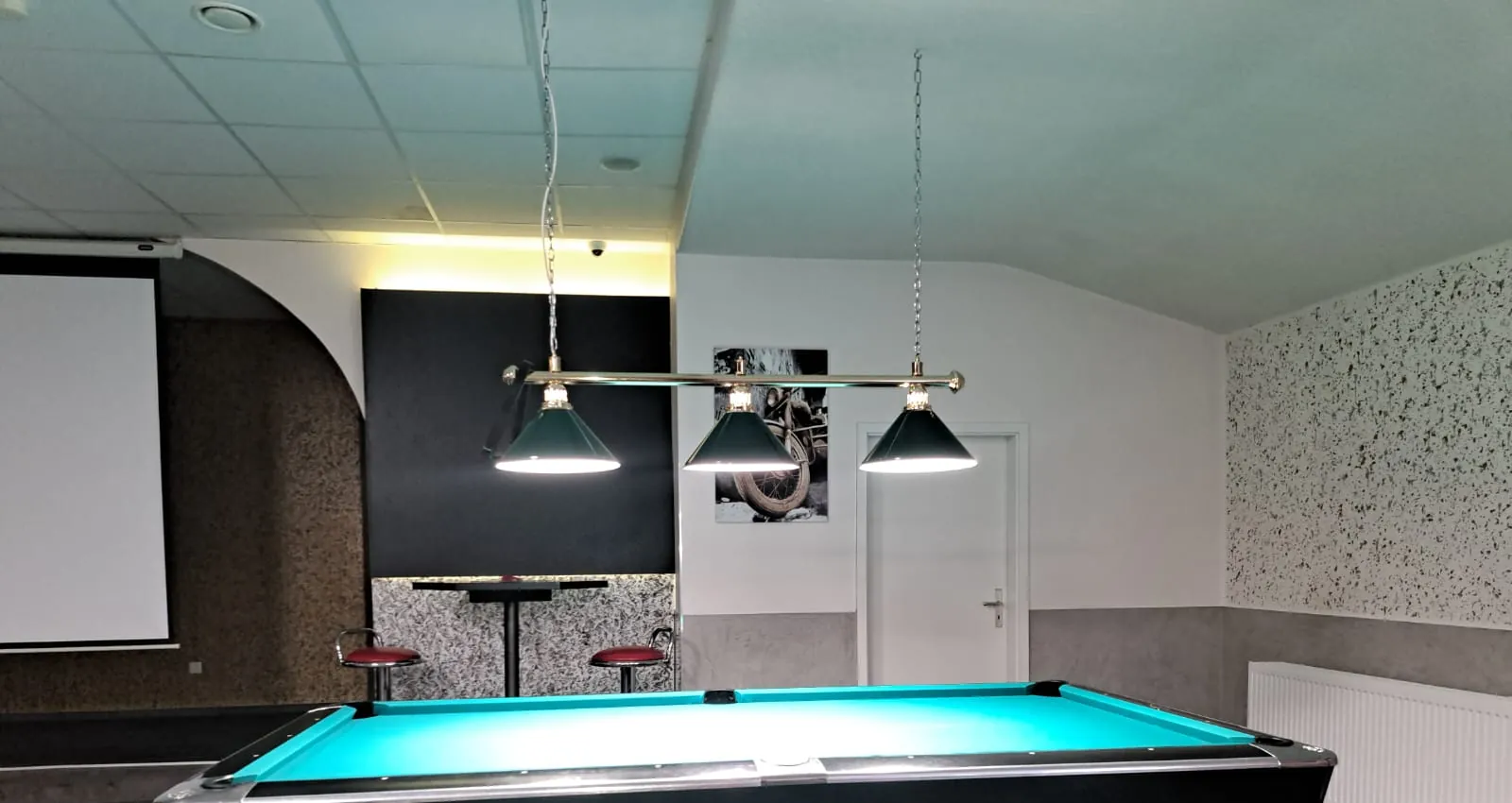 Drei grüne Billardlampen an einer Messingstange über einem Pooltisch bei Billard Wesel, links eine ausgefahrene Leinwand