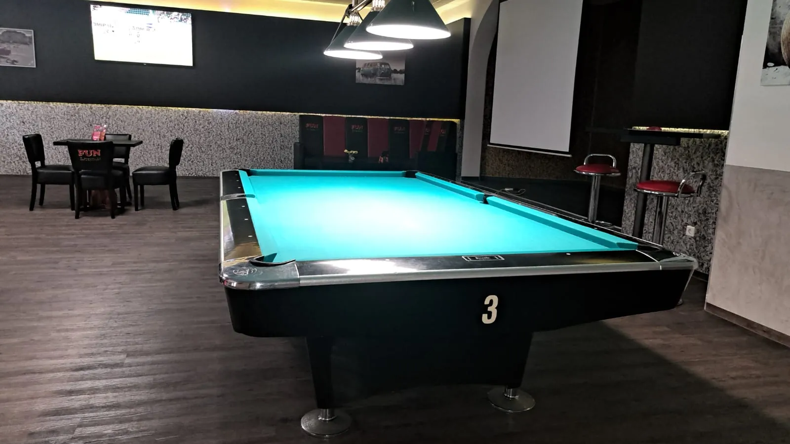 Pooltisch mit türkisfarbenem Tuch und der Nummer 3 unter einer dreiarmigen Lampe bei Billard Wesel, dahinter Leinwand und Stehtisch mit roten Barhockern