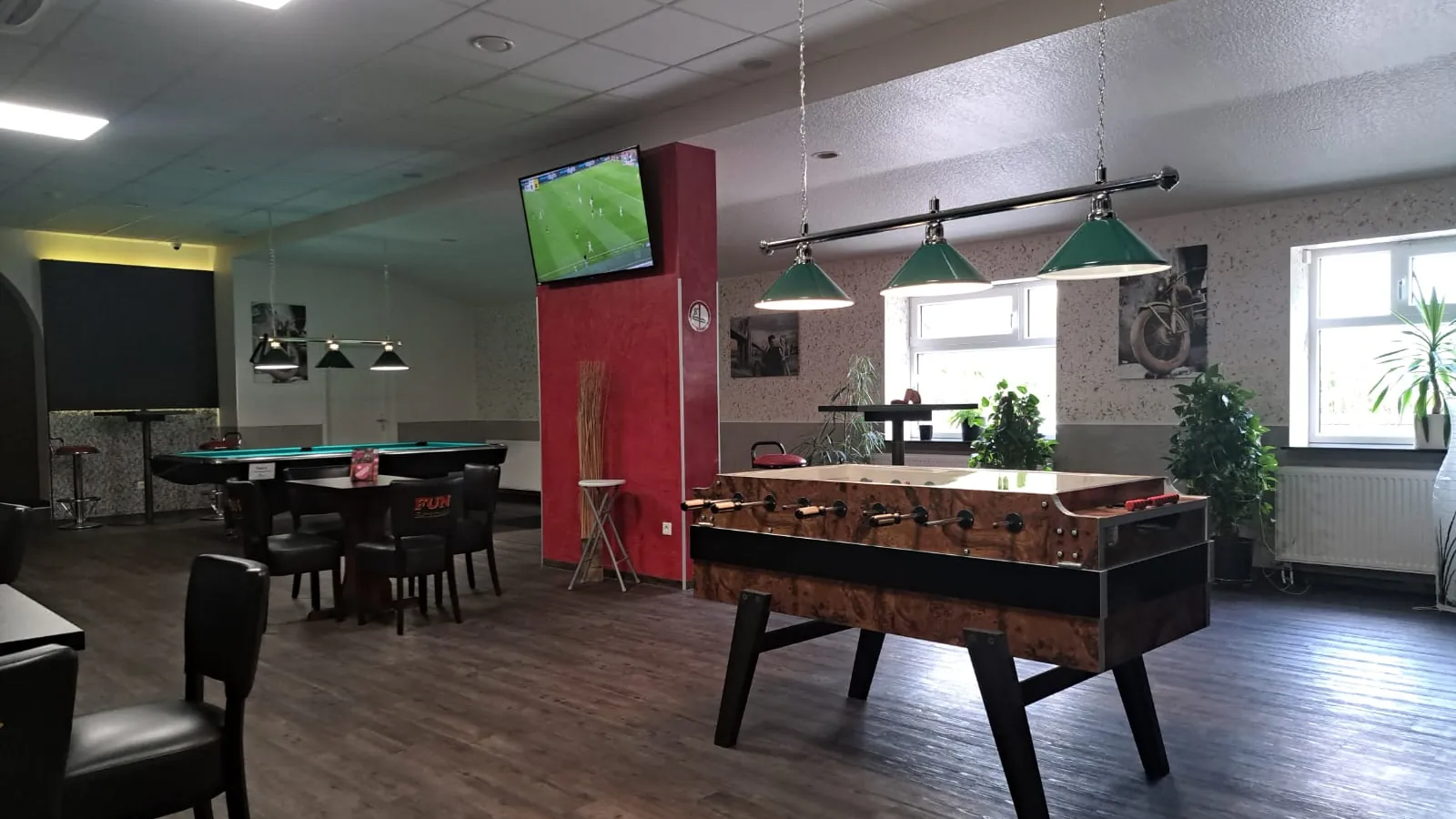 Kicker im Holzdekor unter grünen Billardlampen bei Billard Wesel, dahinter Pooltisch, Fernseher an roter Säule und Zimmerpflanzen am Fenster