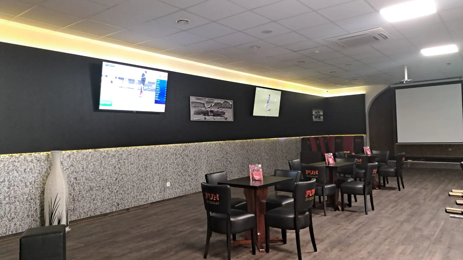 Sitzbereich von Billard Wesel mit schwarzen Lederstühlen an dunklen Tischen, zwei Fernseher an der schwarzen Wand, rechts eine Leinwand
