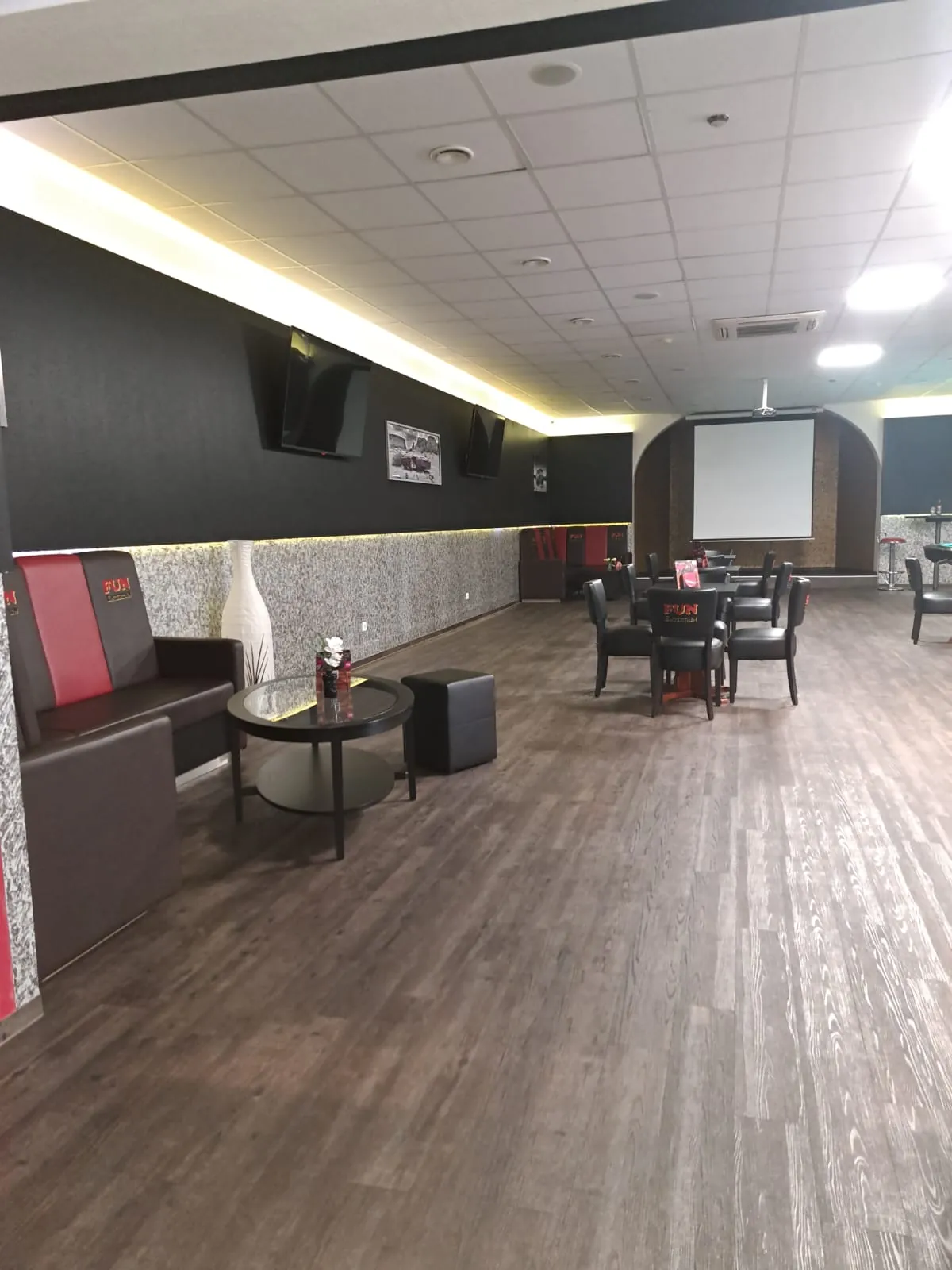 Lounge-Ecke bei Billard Wesel mit rot-schwarzer Sitzbank, rundem Glastisch und Hocker, hinten Tischgruppe und Leinwand